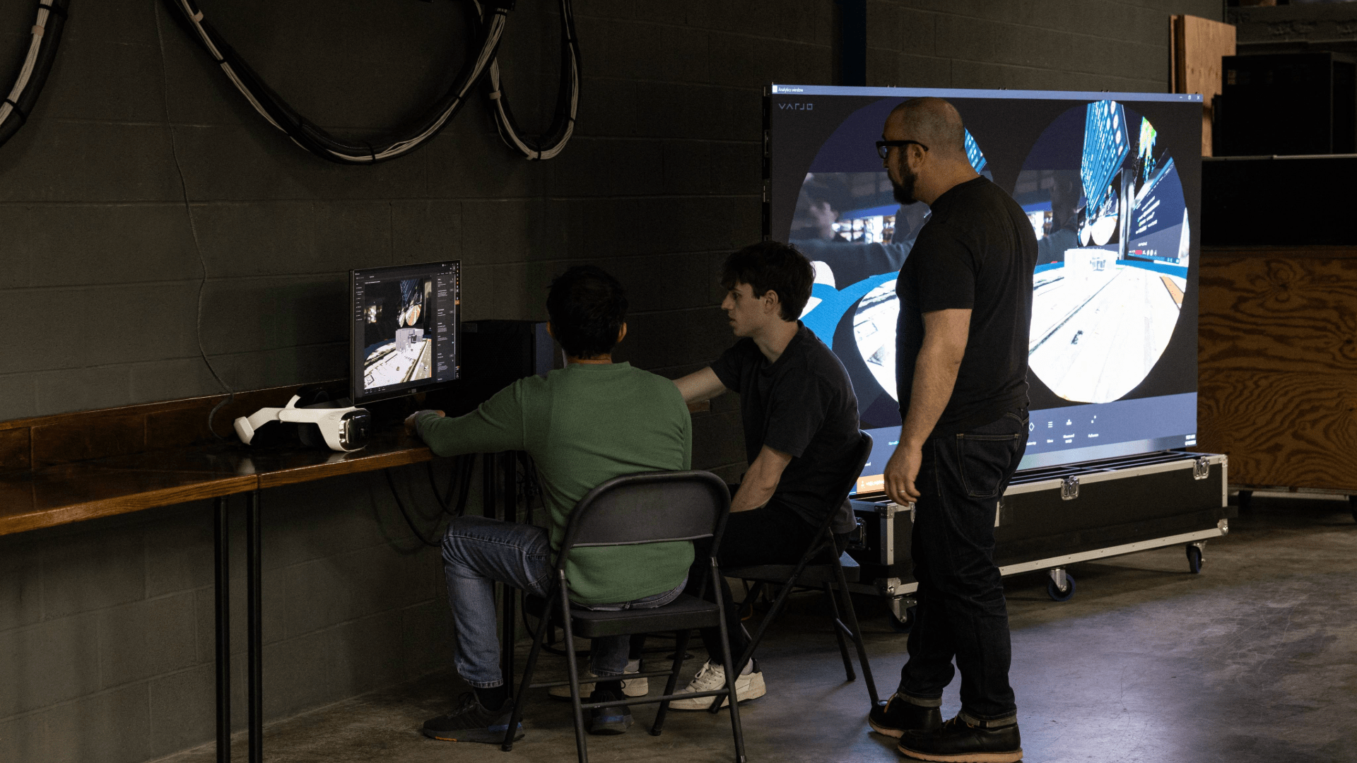 Three people looking at a computer screen, with a bigger screen next to them.