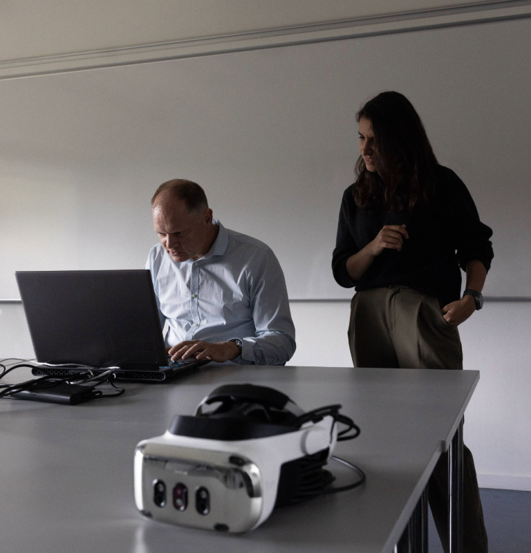 two people looking at a laptop screen, the other one sitting. There's a Varjo XR-4 Series headset on the foreground.
