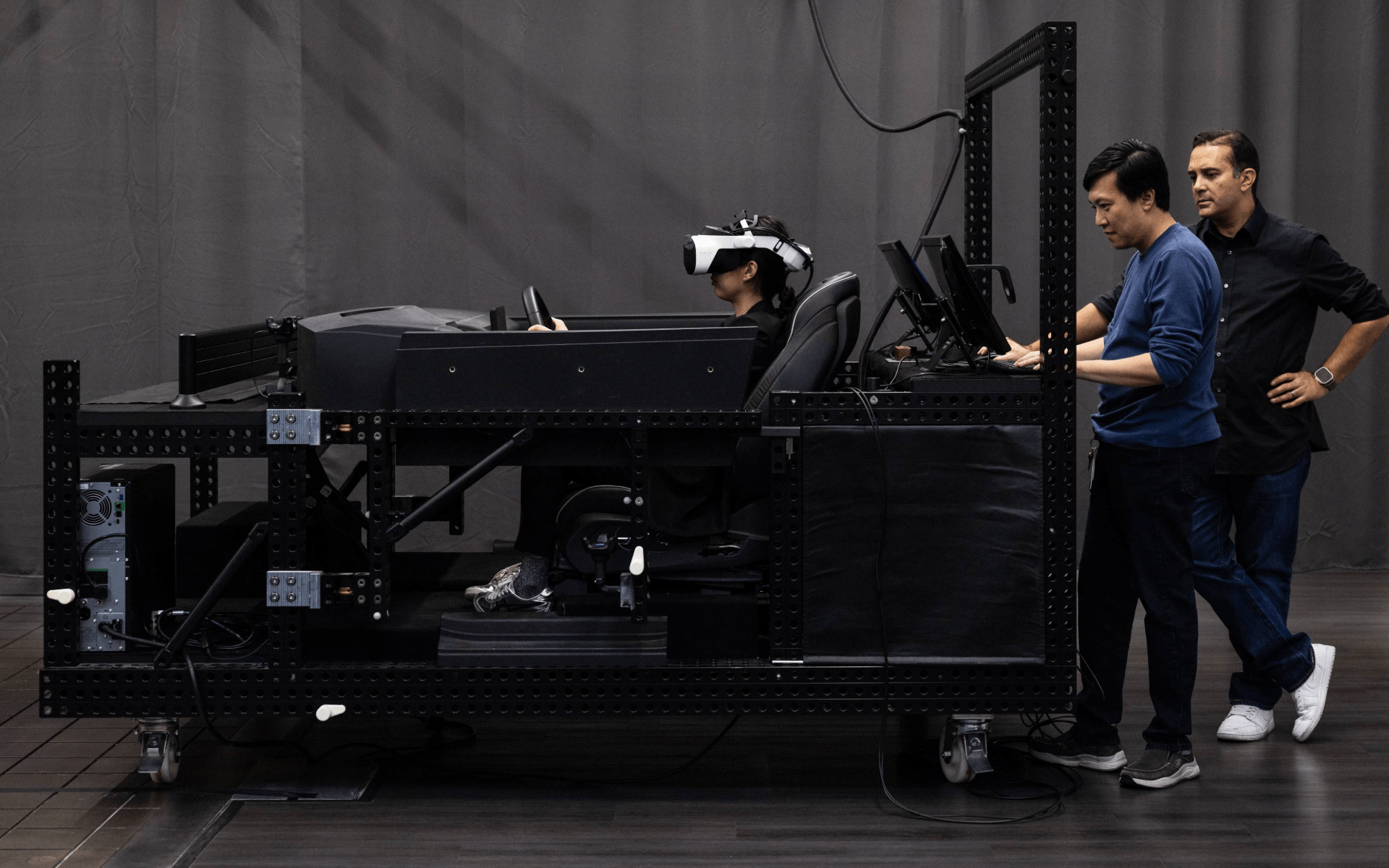 A person wearing a virtual reality headset sits in a black driving simulator rig, gripping the steering wheel as two engineers stand beside the setup monitoring computer screens.